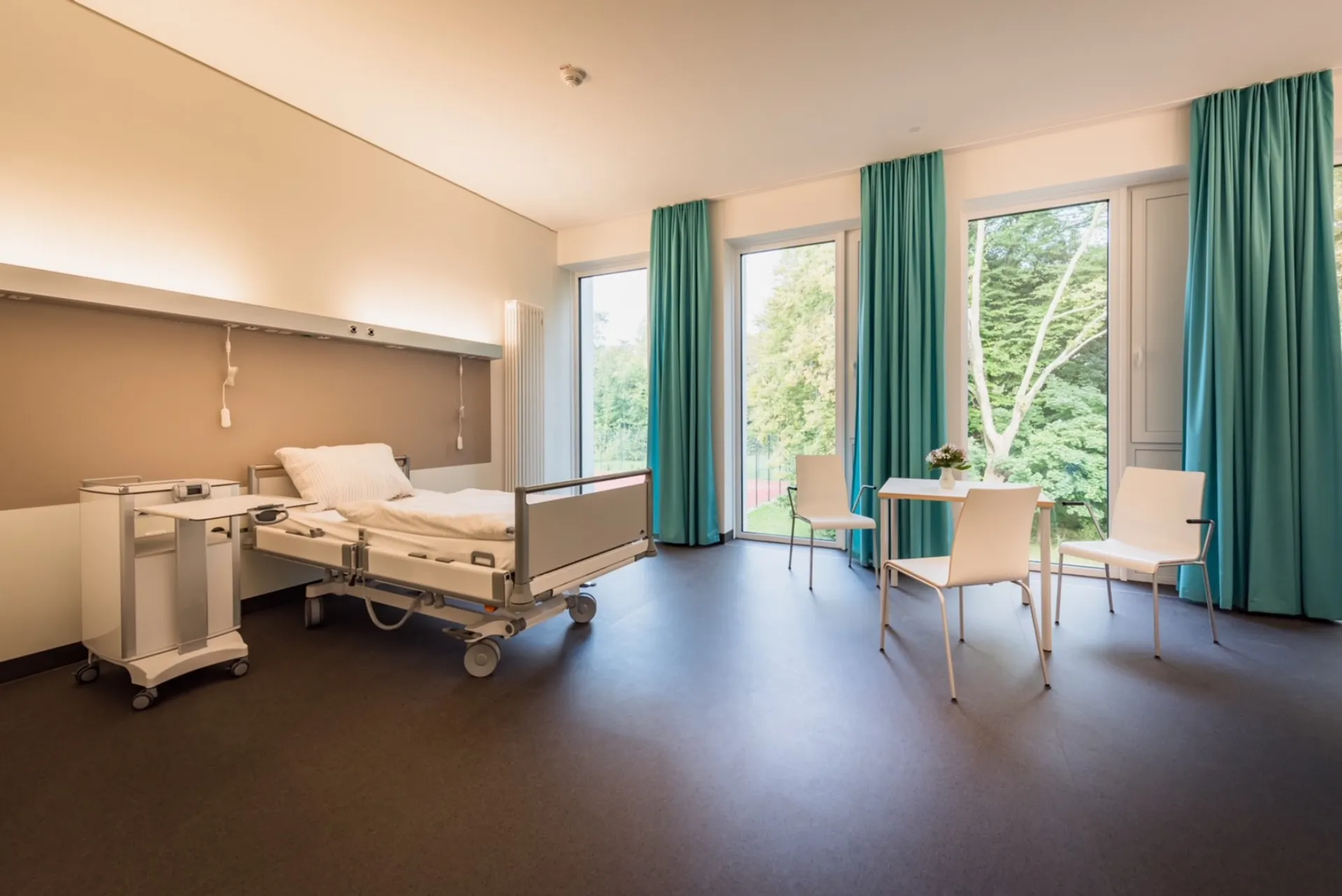 Hospital room with a care bed, a bedside table, a table and chairs in front of large windows with green curtains